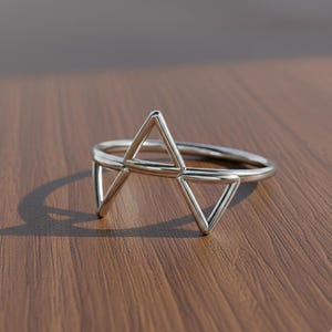 Könnte beinhalten: Ein silberner Ring mit einem geometrischen Design mit drei verbundenen Dreiecken. Der Ring besteht aus glänzendem Metall und liegt auf einer Holzoberfläche. Das Design des Rings ist modern und minimalistisch.