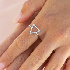 Puede incluir: Un anillo de plata con un diseño minimalista de triángulo abierto. El anillo se lleva en un dedo, mostrando su estética delicada y moderna. La forma geométrica simple del anillo añade un toque de estilo contemporáneo a la joyería.
