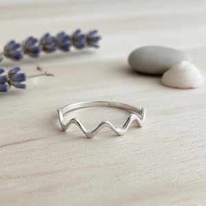 Può includere: Un anello color argento con un design a onda. L'anello è su una superficie di legno chiaro, con rametti di lavanda, una pietra grigia e una conchiglia bianca sullo sfondo. Il design dell'anello ricorda una serie di piccole vette.