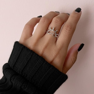 Könnte beinhalten: Ein silberner Ring mit einem Sonnen- und Wellendesign. Der Ring wird an einer Hand mit schwarzem Nagellack und einem schwarzen Ärmel im Vordergrund getragen. Das Design des Rings umfasst eine Sonne mit Strahlen und eine Welle.