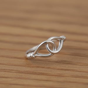 Pode incluir: Um anel de prata com um design de laço duplo. O anel apresenta um acabamento liso e polido e um padrão delicado e entrelaçado. A banda tem uma aparência texturizada perto da base do anel.