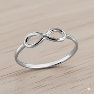 Könnte beinhalten: Ein silberner Ring mit einem Unendlichkeitszeichen. Der Ring ist auf einer hellen Holzoberfläche platziert. Das Unendlichkeitszeichen ist das Hauptmerkmal, mit glatter, polierter Oberfläche.