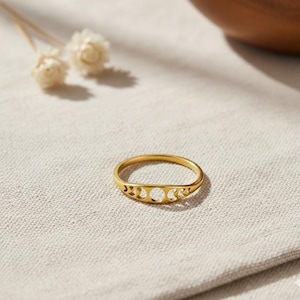 Könnte beinhalten: Ein goldfarbener Ring mit einem Himmelsdesign. Der Ring hat drei Halbmond-Ausschnitte und einen zentralen kreisförmigen Ausschnitt. Der Ring liegt vor einem neutralen Stoffhintergrund, mit weißen Blumen und einer Holzschale im Hintergrund.