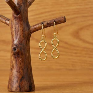 Könnte beinhalten: Ein Paar goldfarbene Ohrringe mit Unendlichkeitszeichen, die an einer Schmuckausstellung aus Holz hängen. Die Ohrringe haben ein schlichtes, elegantes Design mit Hakenverschluss. Die Holz-Ausstellung ist wie ein kleiner Baum geformt.