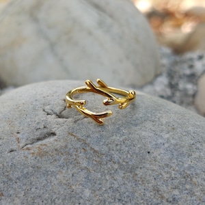 May include: A gold ring featuring a branch design. The ring's open design has branch ends curving inwards. The ring is set against a grey stone background.