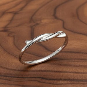 May include: A silver ring with a twisted design. The ring has a smooth, polished finish and sits on a wooden surface. The ring's design features two intertwined bands that create a delicate, elegant look. The ring is a piece of jewellery.