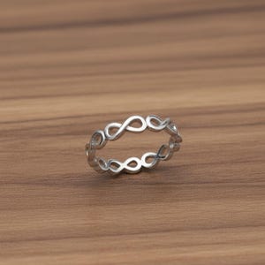 May include: A silver ring with a repeating infinity symbol pattern. The ring is displayed on a warm, brown wooden surface. The infinity symbols are linked, creating a continuous band.