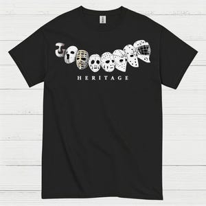 May include: Black t-shirt featuring a series of hockey goalie masks in a row, with the word "HERITAGE" printed below. The masks are in various styles and colors, creating a vintage aesthetic.