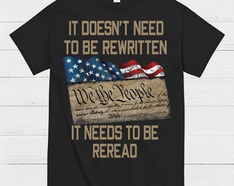 May include: Black t-shirt with the text "IT DOESN'T NEED TO BE REWRITTEN IT NEEDS TO BE REREAD." The design includes an American flag and the words "We the People" in a vintage style.