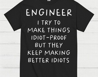 May include: Black t-shirt with white text that reads "ENGINEER I TRY TO MAKE THINGS IDIOT-PROOF BUT THEY KEEP MAKING BETTER IDIOTS." The shirt has a crew neck and short sleeves. The text is in a bold, sans-serif font.