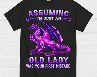 May include: Black t-shirt with a purple dragon graphic. The dragon has large wings and is in a sitting position. Text on the shirt reads "Assuming I'm Just An Old Lady Was Your First Mistake."