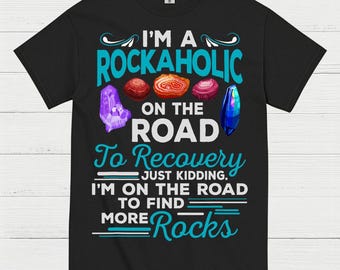 May include: Black t-shirt with the text "I'M A ROCKAHOLIC ON THE ROAD To Recovery. JUST KIDDING. I'M ON THE ROAD TO FIND MORE Rocks." The shirt features colorful illustrations of crystals and gemstones.
