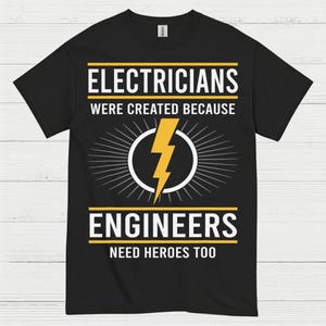 May include: Black t-shirt with the text "ELECTRICIANS WERE CREATED BECAUSE ENGINEERS NEED HEROES TOO". The design includes a yellow lightning bolt inside a white circle, with radiating lines.