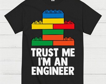 May include: Black t-shirt featuring a colorful graphic of stacked toy building blocks. The blocks are blue, yellow, red, green, and orange. The text "TRUST ME I'M AN ENGINEER" is printed in white below the graphic.