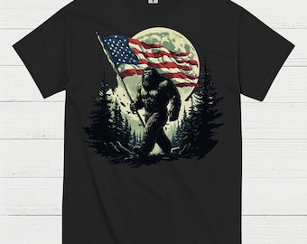 May include: Black t-shirt featuring a Bigfoot graphic carrying an American flag against a full moon backdrop. The design includes pine trees and a detailed illustration of the mythical creature. The flag displays red and white stripes and blue stars.
