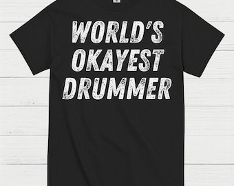 May include: Black t-shirt with the text "WORLD'S OKAYEST DRUMMER" in distressed white lettering. The shirt is a crew neck style and appears to be made of a soft material. The text is centered on the front of the shirt.