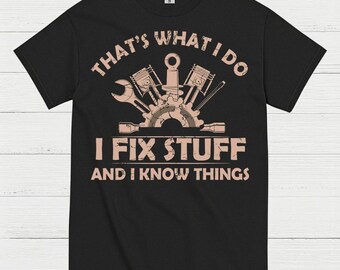 May include: Black t-shirt with a mechanic-themed graphic. The design includes a wrench, pistons, and the text "THAT'S WHAT I DO, I FIX STUFF, AND I KNOW THINGS" in a light brown color. The shirt is made of a soft material.
