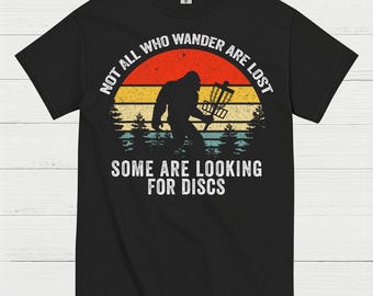 May include: Black t-shirt with a Bigfoot silhouette holding a disc golf basket. The design features a sunset background with the text "NOT ALL WHO WANDER ARE LOST" and "SOME ARE LOOKING FOR DISCS" in white.
