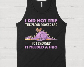May include: Black tank top with the text "I DID NOT TRIP THE FLOOR LOOKED SAD SO I THOUGHT IT NEEDED A HUG." A purple dragon graphic is featured below the text, with orange and yellow accents.