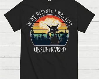 May include: Black t-shirt with a circular graphic featuring a black cat silhouette, a sunset, and the text "IN MY DEFENSE I WAS LEFT UNSUPERVISED". The design includes a forest silhouette and a retro color scheme.