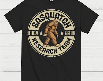 May include: Black t-shirt featuring a circular design with the text "SASQUATCH OFFICIAL BIGFOOT RESEARCH TEAM." The design includes a brown Bigfoot illustration walking, with two pine trees. The shirt is made of a soft material.