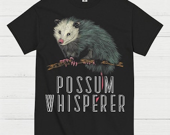 May include: Black t-shirt featuring a detailed illustration of an opossum perched on a branch. The opossum has gray and teal fur, pink nose and paws. The text "POSSUM WHISPERER" is printed below the opossum in white.