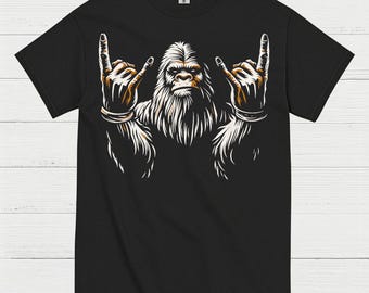 May include: Black t-shirt featuring a white and orange illustration of a Bigfoot-like creature making a rock and roll hand gesture. The design includes detailed fur and facial features, creating a bold graphic.