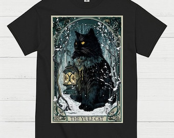 May include: Black t-shirt featuring a detailed illustration of a black cat with yellow eyes holding a lantern in a snowy forest scene. The text "THE YULE CAT" is at the bottom of the design. The shirt is made of a soft material.