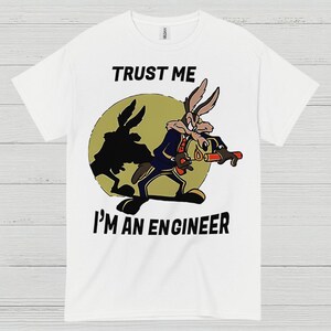 May include: White t-shirt featuring a cartoon image of Wile E. Coyote with the text "TRUST ME I'M AN ENGINEER". The coyote is depicted in a blue suit holding a red and black device, set against a yellow circle.