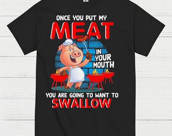 May include: Black t-shirt with a humorous graphic featuring a cartoon pig chef holding a sausage. The text reads "ONCE YOU PUT MY MEAT IN YOUR MOUTH YOU ARE GOING TO WANT TO SWALLOW". Two red grills are also shown.