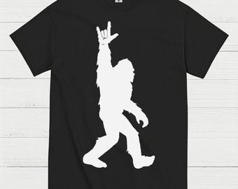 May include: Black t-shirt featuring a white silhouette of a Bigfoot creature making a rock and roll hand gesture. The design is a simple, bold graphic, perfect for fans of cryptids and the outdoors.