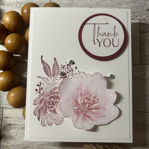 May include: A white card with a burgundy circle that says "Thank You" in white lettering. The card has a pink and burgundy floral design with silver glitter accents.