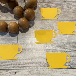 May include: A collection of bright yellow teacup cutouts arranged on a light wood-grain surface. A strand of large, round, wooden beads is in the upper left corner. The teacups are in various orientations.