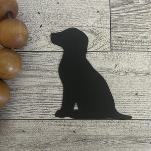 May include: Black metal silhouette of a dog sitting with its head turned to the side. The silhouette is on a light wood background.