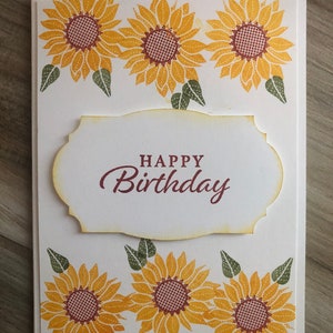 May include: A white card with a brown label that says "Happy Birthday" surrounded by yellow sunflowers with brown centers and green leaves.
