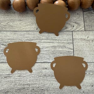 May include: Three brown cauldron cutouts with handles and legs are arranged on a light wood-grain surface. Wooden beads and twine are in the background. The cutouts are for a Halloween or witch-themed craft.