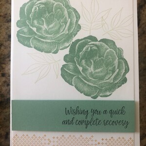 May include: A white card with two large sage green rose illustrations and delicate leaf outlines. A sage green band across the center reads "Wishing you a quick and complete recovery." A gold patterned border is at the bottom.
