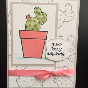 May include: A greeting card with a cactus illustration and the text "make today amazing". The card has a pink ribbon tied around it.