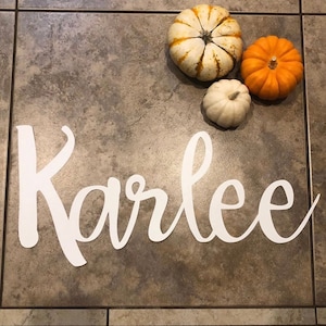 May include: White cursive script spelling the name "Karlee" on a brown tiled floor with three small pumpkins, one white and two orange.