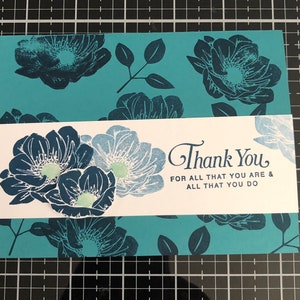 May include: A teal card with a white border and a floral design. The card has the text "Thank You" with the message "For all that you are & all that you do".