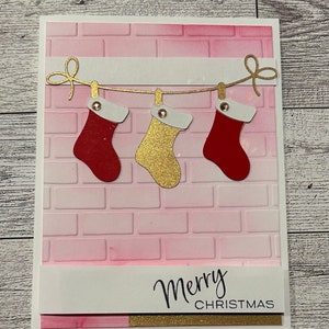 May include: A pink and white Christmas card with a brick pattern background. Three stockings are hanging from a string with gold bows. The stockings are red, gold, and red. The card says "Merry Christmas" in black script.
