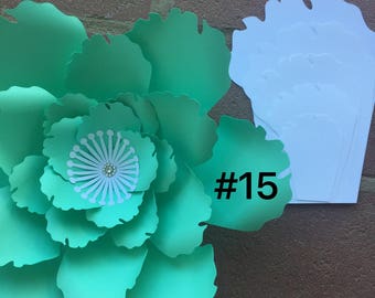May include: A large, handmade paper flower in shades of green, with a white and silver center. The flower is set against a brick wall. A stack of white paper petals is to the right, with the number "#15" visible.
