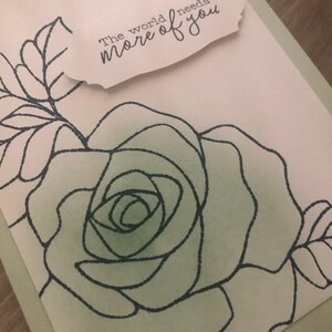 May include: A greeting card with a green and white watercolor background featuring a black line drawing of a rose. The card has a white tag with the text "The world needs more of you" written in black.