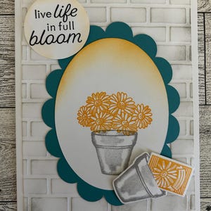 May include: A handmade greeting card with a white brick background. The card features a teal scalloped oval with a bouquet of orange daisies in a gray pot. The text "live life in full bloom" is in a circle at the top.