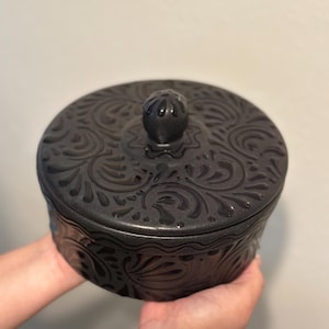 May include: A round, black ceramic container with a lid. The container and lid are adorned with an embossed floral pattern. The lid features a small, round knob for easy opening. The container is held in front of a neutral background.