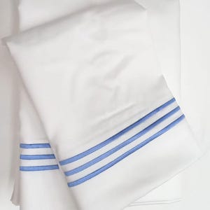 Puede incluir: Sábanas blancas con tres rayas paralelas bordadas en azul claro a lo largo del borde. Las sábanas están dobladas y apiladas, mostrando el diseño. La ropa de cama es de un material liso, probablemente algodón.
