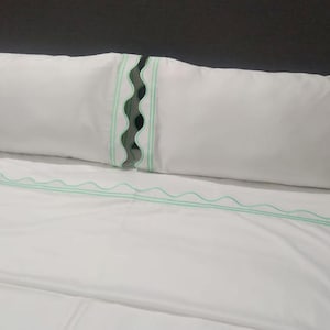 May include: White bed linens with a decorative mint green scalloped edge on the pillowcase and a matching wavy trim on the sheet. The pillow is long and rectangular, resting on the bed.