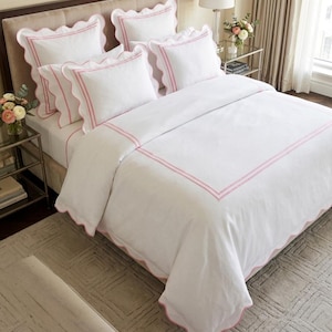 May include: A white bedding set featuring a duvet cover and multiple pillow shams with scalloped edges and double pink embroidered borders. The set includes a large duvet cover and four rectangular pillows, all displaying a matching pink trim.