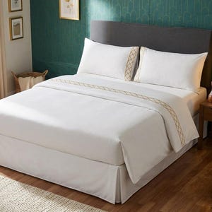 Puede incluir: Un juego de cama blanco que incluye una funda nórdica y dos fundas de almohada a juego, decoradas con un borde geométrico de color dorado. La ropa de cama está dispuesta sobre una cama con cabecero gris oscuro y faldón blanco.