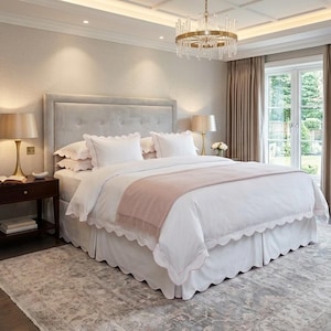 May include: A king-sized bed featuring a tufted light gray headboard, white bedding with scalloped edges, and a soft pink throw blanket. The bed is flanked by two dark wood nightstands with gold lamps, set against a neutral-toned bedroom interior.